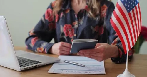 Mujer sosteniendo un pasaporte en un escritorio con un formulario de solicitud de visa y una pequeña bandera estadounidense.