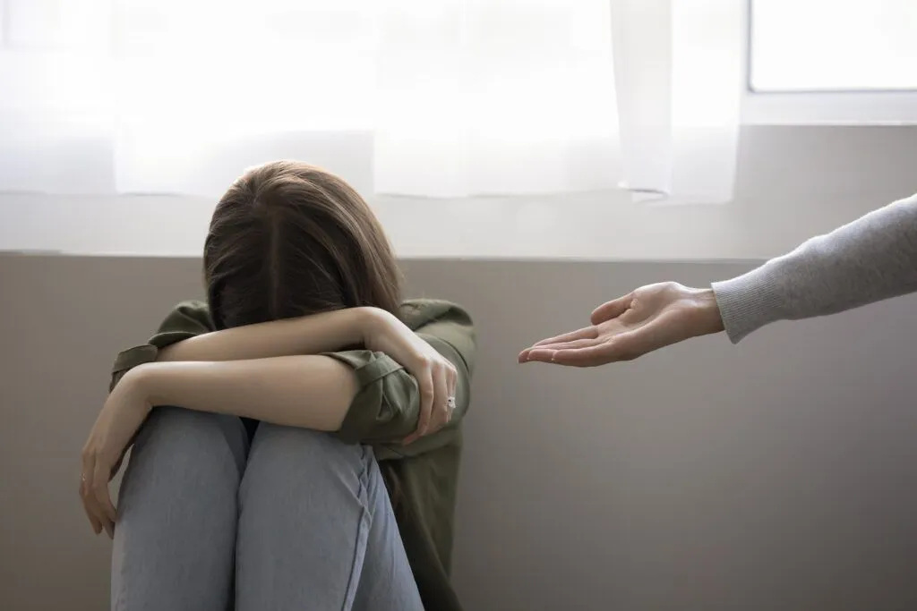 Una joven sentada en el suelo con la cabeza agachada y los brazos cruzados sobre las rodillas, mientras otra persona le tiende la mano en señal de apoyo.