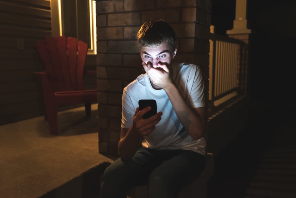 Un adolescente reacciona a algo impactante en su teléfono mientras está sentado fuera de su casa por la noche.