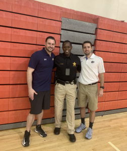 Adam and Manny from Rossen Law Firm standing on a basketball court with PBSO Deputy Kenneth 'Coach T' Torrence and students at the Tri-City Basketball, Academics and Mentoring Camp