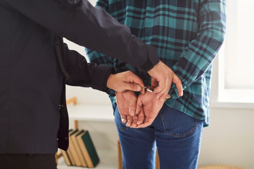 Cropped image of a male police officer arresting a handcuffed man inside a home, suspected of committing a crime.