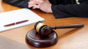 Close-up of a judge's gavel on a wooden table. A woman sits with folded hands and a notepad in front of her, symbolizing justice or auction proceedings.